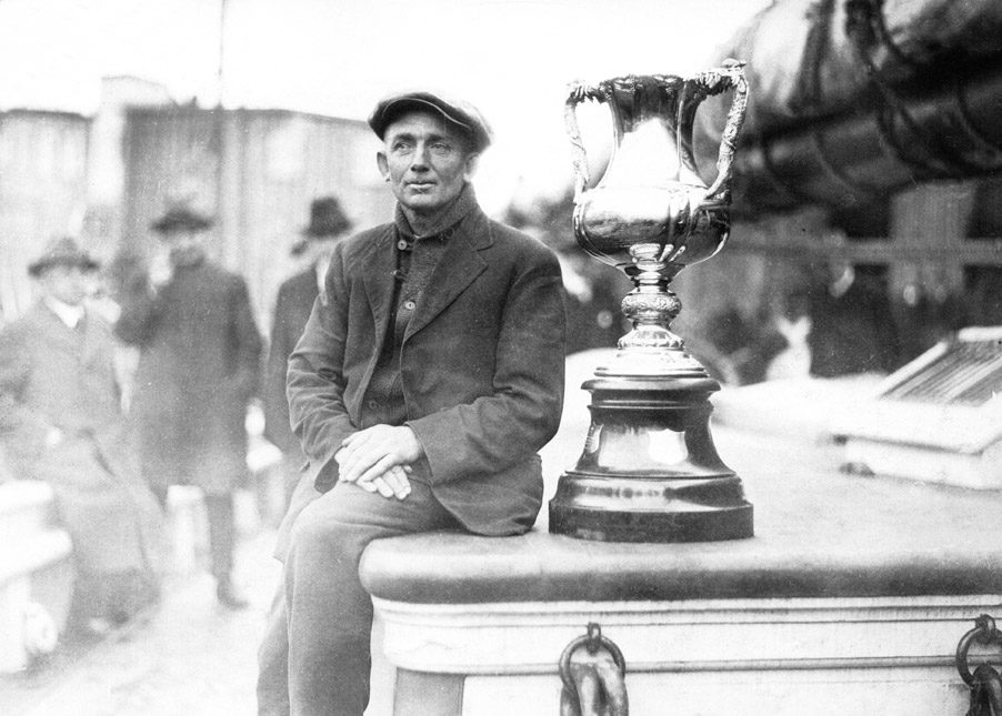 Captain Angus Walters seated on the deck of the Bluenose beside the tall silver International Fishermen's Trophy, 1921.