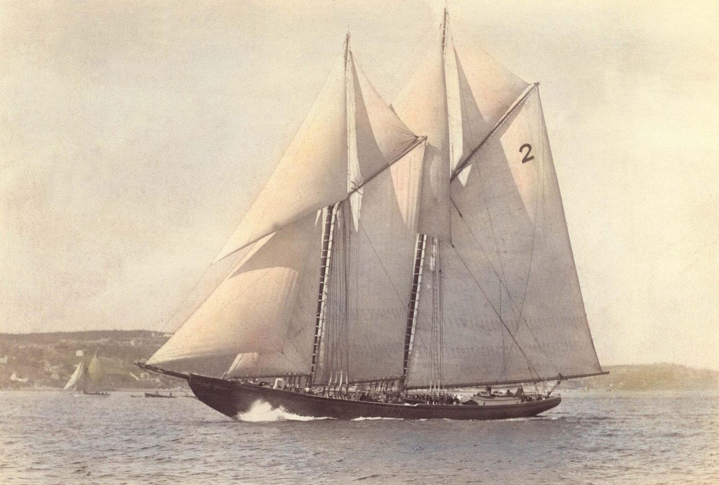 The Bluenose under full sail off Halifax in October 1921, a two-masted schooner with a large racing number 2 on her mainsail, heeling slightly, with a low shore behind her.