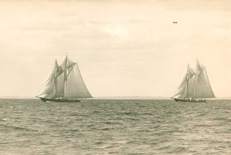 Two gaff-rigged fishing schooners racing close together off Gloucester in 1922, a seaplane overhead.