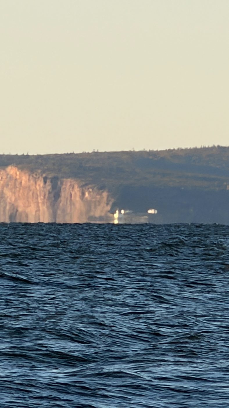 Cape d'Or seen across choppy water from the far shore of Minas Channel: red cliffs, the white light station on the point, the hills behind.