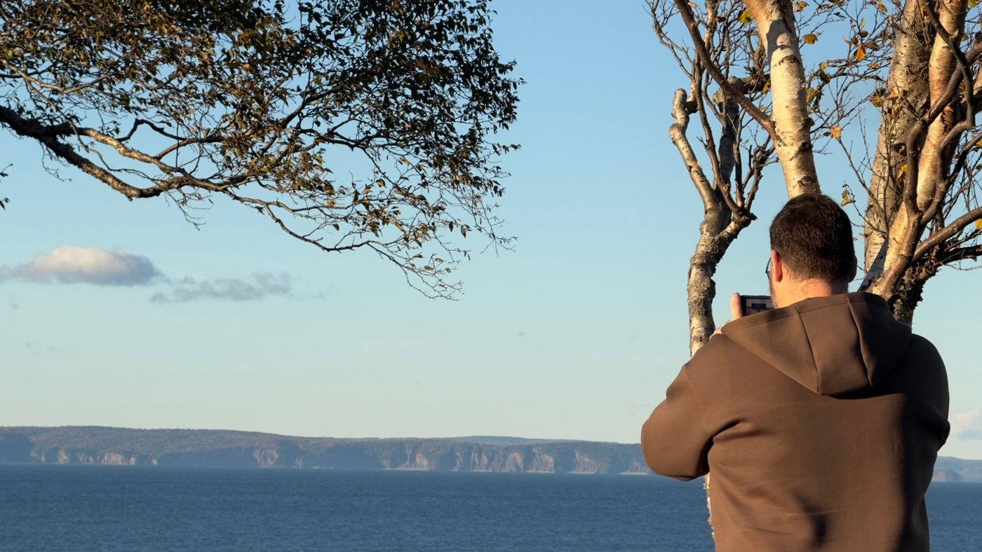 A man in a brown hooded jacket, seen from behind under a tree, films the far shore of Minas Channel with a phone; Cape d'Or is a long line of cliffs across the water.