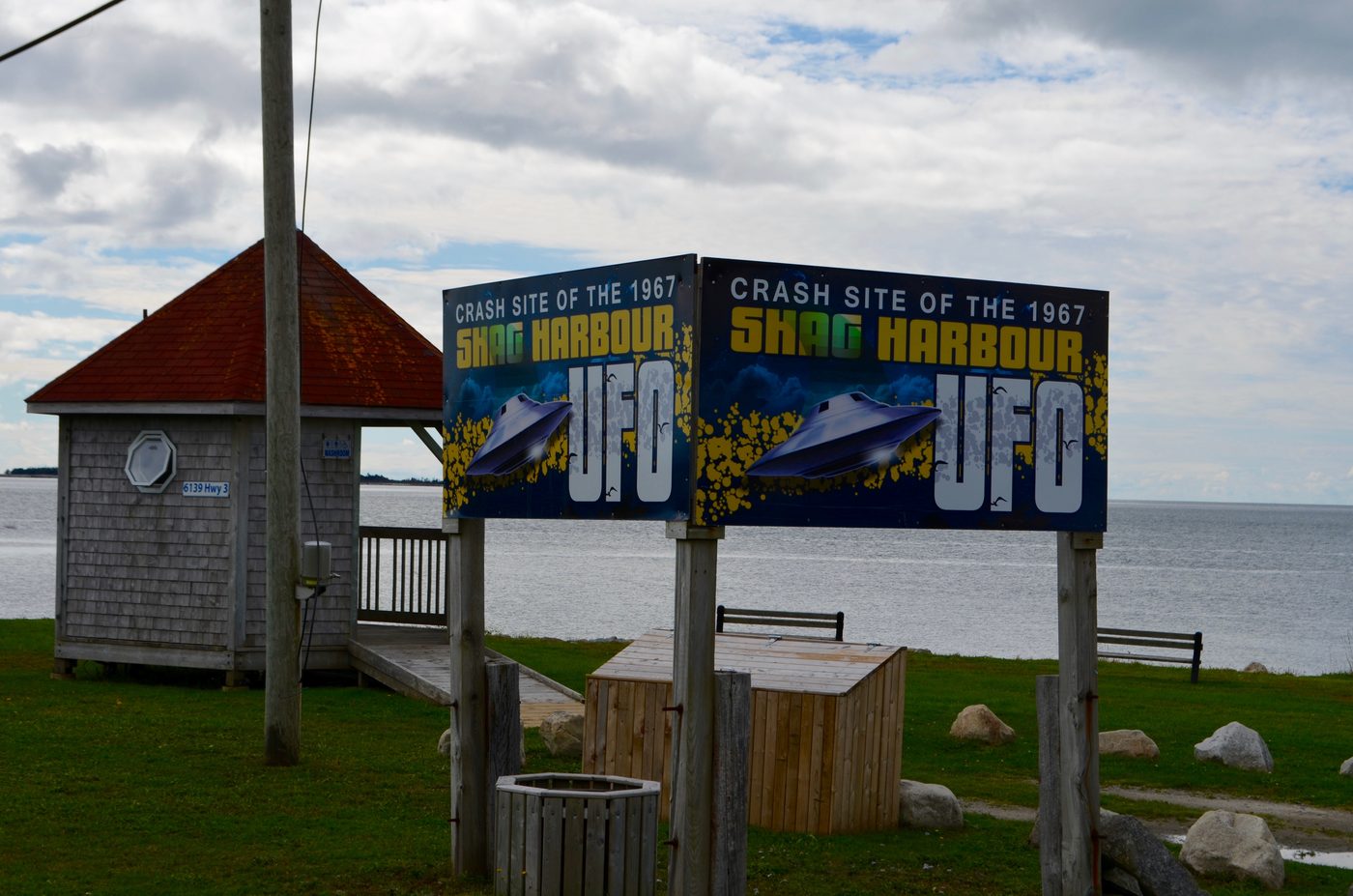 A shingled gazebo with a red roof beside two painted signs reading Crash Site of the 1967 Shag Harbour UFO, on a lawn above the sea.